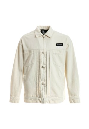 Joven de pie con chaqueta crema, pantalones blancos, zapatos marrones claros y una camiseta blanca con un dibujo de un oso de peluche sobre un fondo blanco.