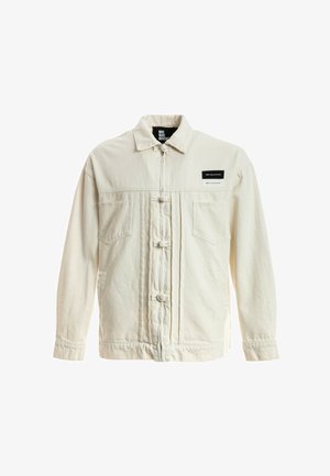 Joven de pie con chaqueta crema, pantalones blancos, zapatos marrones claros y una camiseta blanca con un dibujo de un oso de peluche sobre un fondo blanco.