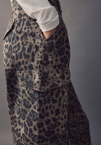 Jeanslatzhose mit Leopardenmuster und Seitentasche, mit einem gedämpften braunen Grundton und schwarzen Flecken, die eine strukturierte Oberfläche zeigen.