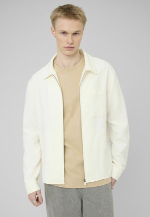 Jeune homme portant une veste écrue dézippée sur une chemise beige et un pantalon gris, debout devant un fond uni gris clair.