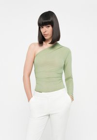 Asymmetrische top met één schouder in helder lichtgroen, met subtiele plooien en een aansluitende silhouet, gecombineerd met witte, op maat gemaakte broek.