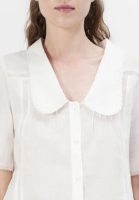 Hvit linbluse med en stor, rund krage, korte ermer og detaljerte blondeakenter langs sømmene. Har perlelignende knapper.