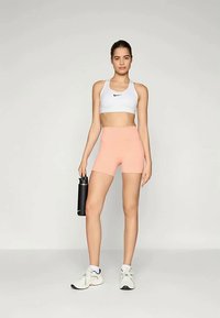 Fehér sportmelltartó fekete logóval, rózsaszín sportnadrág, és fekete vízpalack. A modell fehér tornacipőt visel, és egy sima háttér előtt áll.