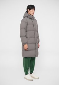 Šedý prešívaný kabát s kapucňou, s hrubým výplňom, dlhým strihom a prednými vreckami, skombinovaný so zelenými nohavicami a bielymi sneakers.