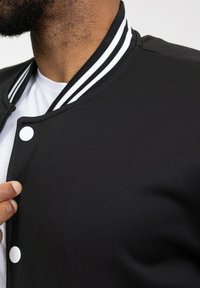 Veste de varsity noire avec col à côtes blanches et boutons pressions blancs. Le tissu semble lisse, s'ajustant de près au cou.