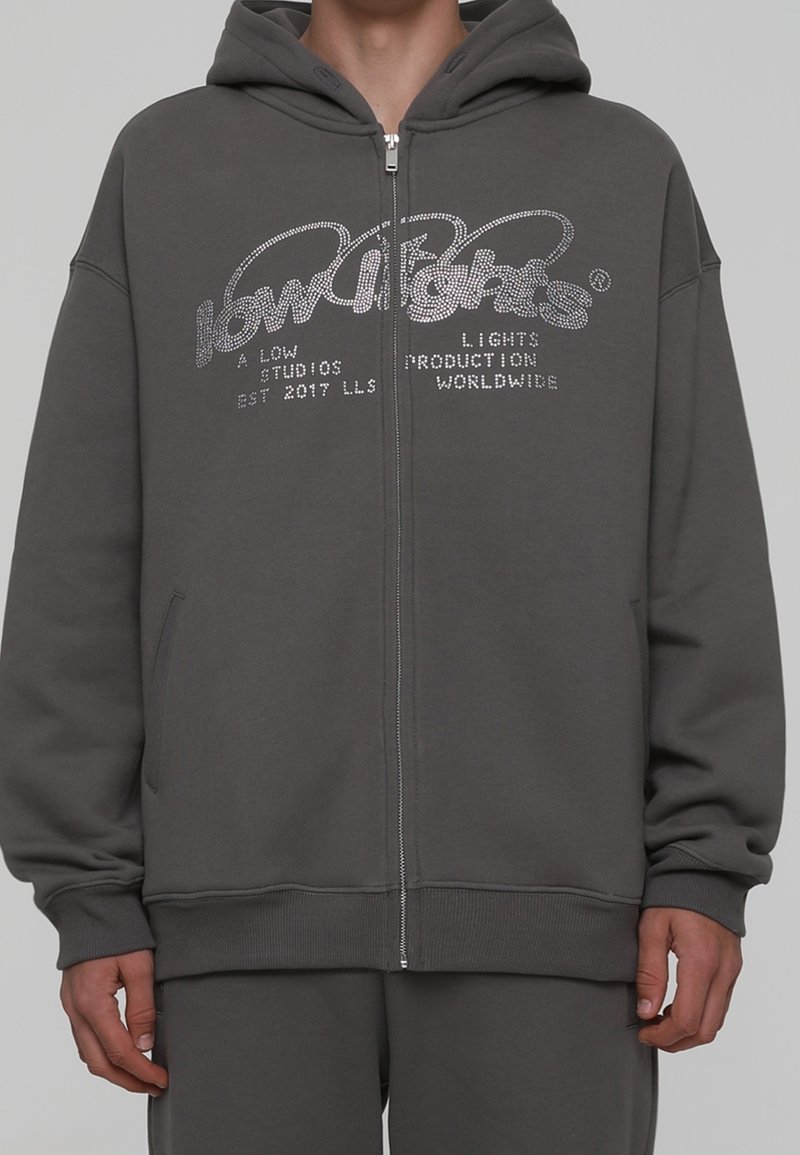 Sudadera con cremallera en gris, con capucha ajustable y gráficos impresos en gris claro. Está hecha de una tela suave que muestra un corte relajado y bolsillos laterales.
