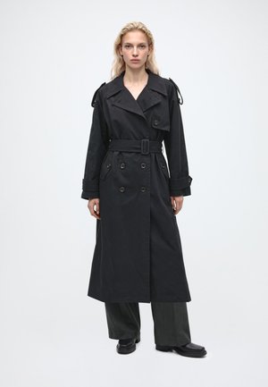 Femme aux cheveux blonds portant un long trench-coat noir ceinturé, un pantalon large foncé et des chaussures noires, debout devant un fond clair uni.