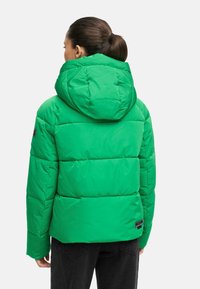 Veste doudoune verte avec capuche, présentant un design matelassé et une épaisse isolation. Détail étiquette noire près de l'ourlet ; texture brillante.