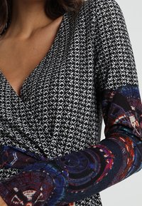 Langärmliges Wickeloberteil mit einem schwarz-weißen geometrischen Muster am Körper und farbenfroh gemusterten Ärmeln. Weicher, dehnbarer Stoff.