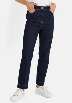 Persoon draagt donkerblauwe jeans met rechte pijpen en omgeslagen zomen, witte casual sneakers en heeft handen in de voorzakken tegen een effen achtergrond.