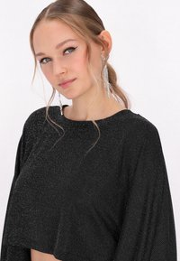 Svart kort topp med en lös passform, som har en texturerad yta med silverprickar. Modellen bär hängande örhängen med kristallaccenter.