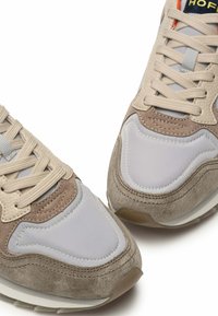 Beige und graue Sneaker mit Wildleder- und Mesh-Oberflächen, ausgestattet mit einer Schnürung, verstärkter Zehenkappe und einem kontrastierenden Logo auf der Zunge.
