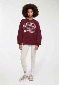 Felpa bordeaux con testo bianco "MANHATTAN New York EAST COAST," abbinata a leggings grigi chiari e sneakers beige con accenti bianchi e grigi.