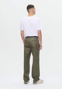 Olivgrüne, weit geschnittene Hose mit glatter Textur, zwei Gesäßtaschen und gesticktem Logo. Kombiniert mit einem schlichten weißen T-Shirt und Sneakers.