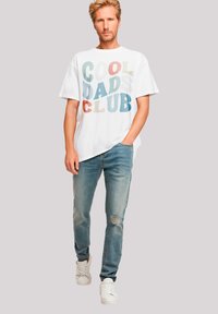 White t-shirt with multicolored "COOL DADS CLUB" text, paired with light blue ripped jeans and white sneakers. Casual fit and lightweight fabric.