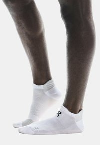 Weiße Sportsocken mit strukturierten Mustern und elastischen Bündchen, die mit schwarzen Logodetails versehen sind und sich eng um die Knöchel der Füße schmiegen.