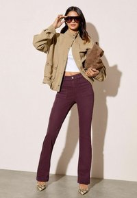 Jachetă supradimensionată khaki, top scurt alb, pantaloni evazați prune cu buzunare frontale, poșetă din suede maro și pantofi cu toc înalt cu model.