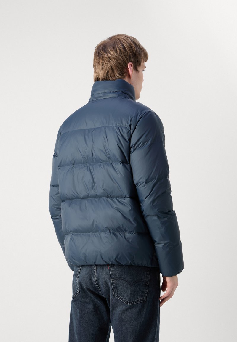 Marineblaue Steppjacke mit hohem Kragen und gestepptem Design. Glatte Textur und taillierte Ärmel, von hinten betrachtet.