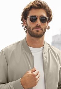 Hellbeige Bomberjacke mit geripptem Kragen und Reißverschluss, getragen über einem weißen T-Shirt. Klare runde Sonnenbrille mit dunklen Gläsern.