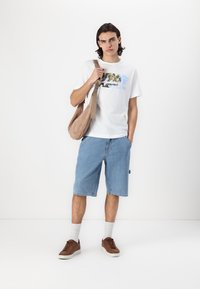 Hvid grafisk t-shirt med en strandscene, blå denimshorts, brune sneakers og en beige skuldertaske. Modellen står mod en lys baggrund.