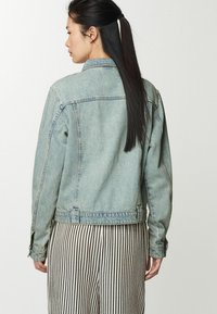 Giacca di jeans blu chiaro con una texture leggermente strappata, dettagli in cucitura e polsini con bottoni, abbinata a pantaloni a righe bianco e neri.