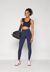 Svart trenings-BH, høy Midje marineblå leggings med sidedetaljer, hvite aksenter og en svart treningsbag. Bært med hvite joggesko.