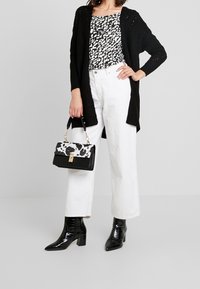 Černý pletený cardigan přes bílou a černou vzorovanou blůzu, doplněný bílými kalhotami širokého střihu a lesklými černými botami na podpatku. Drží černobílou tašku.