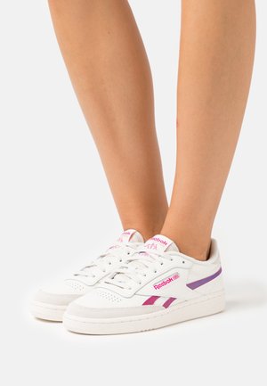 Baskets blanches Reebok avec des détails roses et violets portées pieds nus, posées sur un fond blanc.