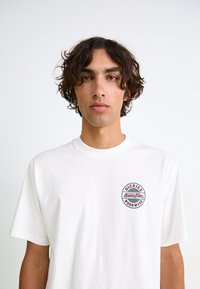 Hvid bomulds T-shirt med et cirkulært rødt og blåt logo på venstre bryst, der står "DICKIES WORKWEAR." Korte ærmer og en afslappet pasform.