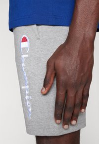 Pantalones cortos de sudadera grises con un logotipo bordado "Champion" en blanco, con un acento rojo y azul. Material suave de mezcla de algodón, ajuste relajado.