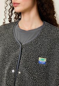 Pelēks, teksturēts vilnas vestes jaciņa ar apaļu kakla izgriezumu, spiesta aizdare un krāsains plāksteris ar uzrakstu "AMV" krūtīs. Valkāta virs pelēkās krekliņa.