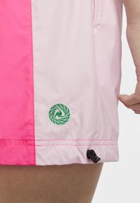 Veste légère avec un schéma de couleurs rose et rose pastel, arborant un logo vert brodé, un ourlet élastique et un rideau de tirage.