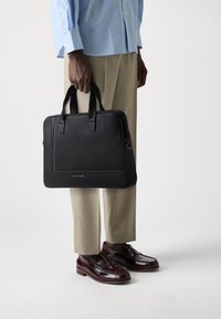 Black textured leather briefcase with dual top handles, a zip closure, and subtle logo detailing on the front. Adjusted for formal wear.