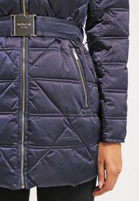 Marineblå quilted puffersjakke med bælte i taljen, der har sølvhardware, lynlåslommer og diagonale stikninger.