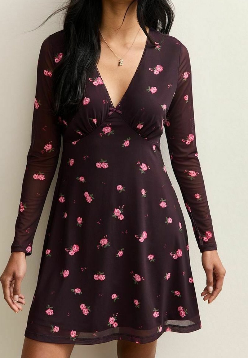 Femme portant une robe longue à manches longues violet foncé avec un motif floral rose et un décolleté en V, debout devant un fond uni.