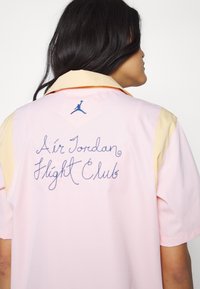 Chemise rose à manches courtes avec un col jaune, présentant un texte et un logo brodés "Air Jordan Flight Club" en bleu à l'arrière.