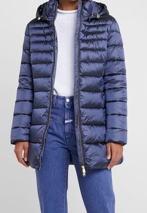 Persoon draagt een marineblauwe doorgestikte donzen jas over een witte gebreide trui en blauwe jeans met hoge taille, staand tegen een effen achtergrond.