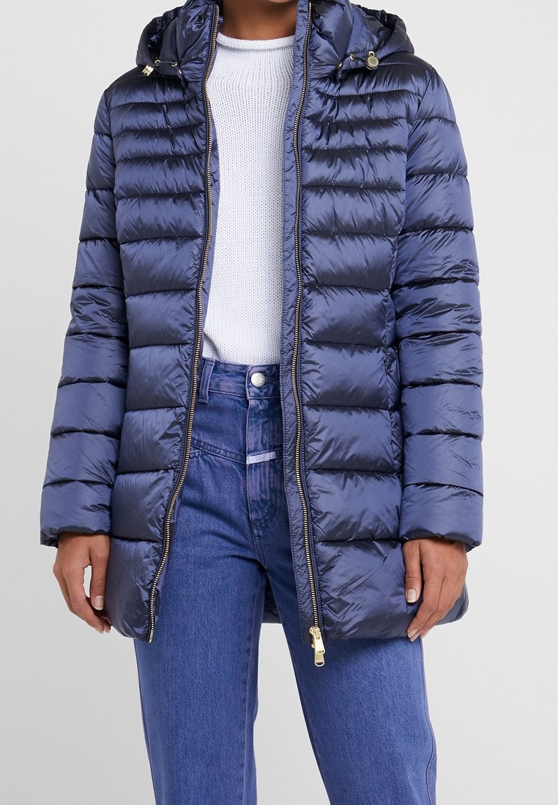 Personne portant une doudoune matelassée bleu marine par-dessus un pull en maille blanc et un jean bleu taille haute, debout devant un fond uni.