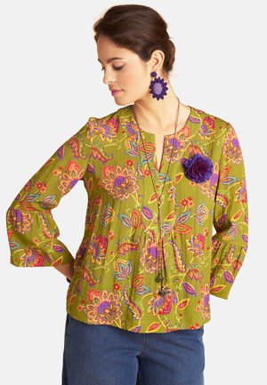 Frau trägt grüne Bluse mit Blumenmuster, einer lila Blumenbrosche, passenden lila Blumenohrringen und langer Halskette, schaut nach unten, Hände in die Hüften gestützt.