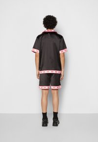 Person with curly hair stands facing away, wearing black satin short-sleeve shirt and shorts with red-and-white patterned trim and black shoes.