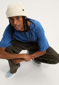 Camiseta de manga corta azul combinada con pantalones holgados verdes oliva y zapatillas slip-on a cuadros azules y blancos. Gorra de punto crema con canalé.