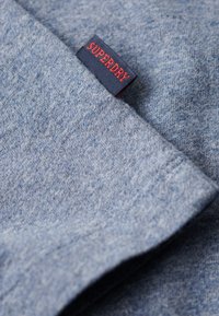 Tissu en coton bleu au toucher doux, avec une étiquette "SUPERDRY" visible en marine et rouge sur le bord.