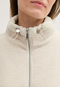 Veste zippée beige avec un col haut texturé crème. Comprend des garnitures en métal argenté et des cordons de réglage au niveau du col.