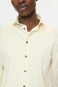 Lichtgeel shirt met een button-down kraag, voorzien van donkerbruine knopen en een zachte, gestructureerde stof. Close-up van de halslijn.