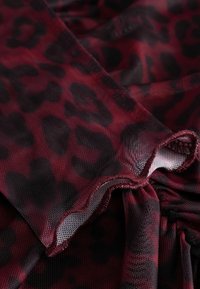 Tkanina w czerwono-czarny wzór panterki z teksturowaną siateczką, z marszczonym brzegiem i gładkim wykończeniem, sugerującym miękkość i elastyczność.
