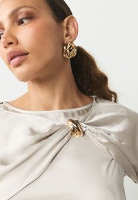 Pendientes de oro con nudos y una blusa de satén gris claro con un diseño torcido y un detalle metálico en el escote. Textura suave.