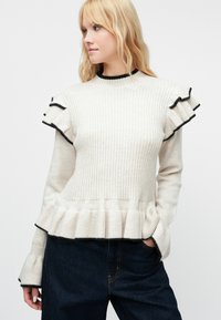 Svetlo bež rebrast pulover s črnimi detajli, z nabranimi rokavi in peplum robom, izdelan iz mehke teksturirane tkanine.