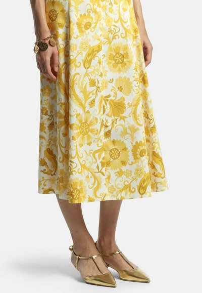 Kvinde iført nederdel i gul blomsterprint, armbånd med guldmønter og gyldne spidse T-strop hæle, stående mod en lyse baggrund.