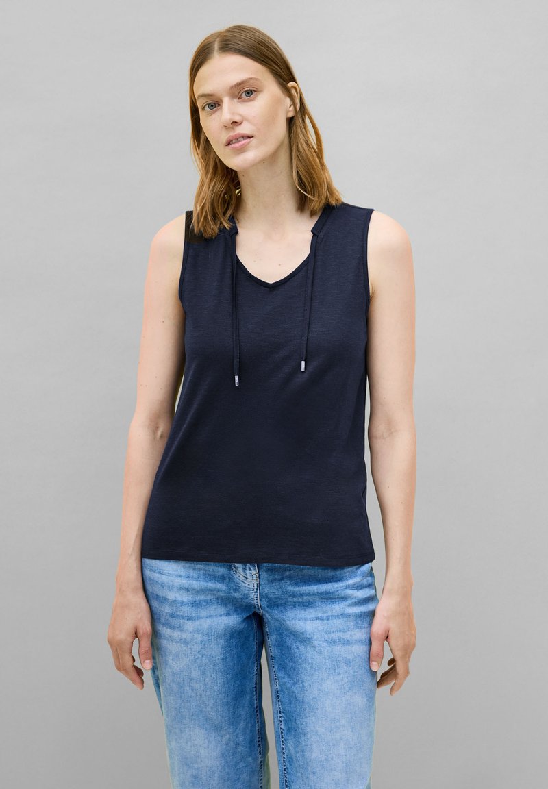 Top senza maniche di colore blu navy con scollatura a V e cordino regolabile. Tessuto morbido e liscio in vestibilità rilassata, abbinato a jeans in denim azzurro chiaro.