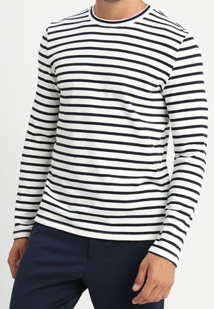 Muž v bílém tričku s dlouhým rukávem s černými vodorovnými pruhy a tmavě modrými kalhotami, stojící před jednobarevným pozadím.